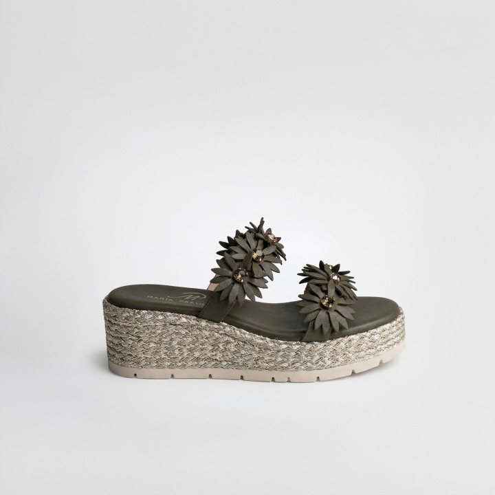 Oliwkowy klapek Maria Pakulnis na koturnie z plecionym wykończeniem. Dwa paski zdobione trójwymiarowymi kwiatami z biżuteryjnym środkiem. Jasna, ząbkowana podeszwa i logo na wkładce. Widok z profilu na białym tle.